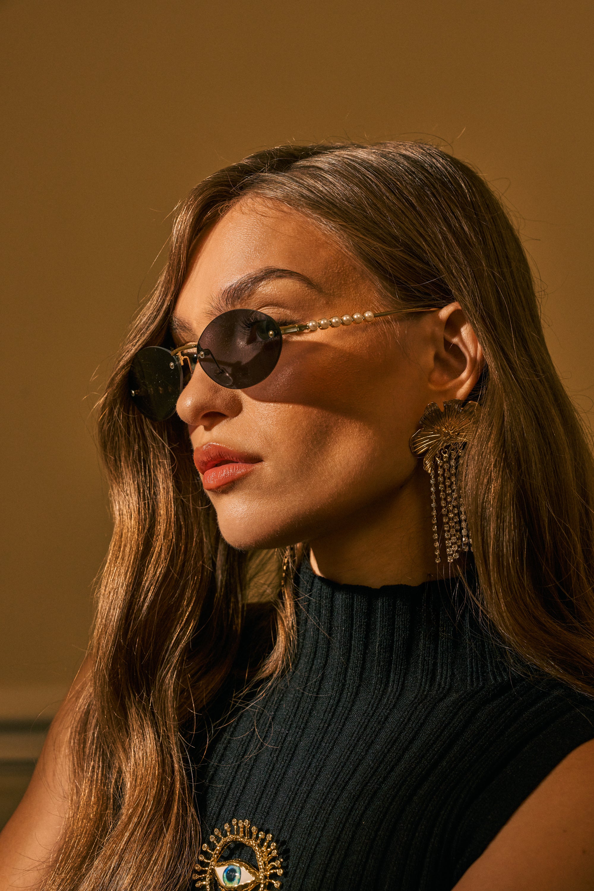 Wearing the SEE YA SUNGLASSES, a woman with long wavy hair, statement earrings, and a sleeveless black top adorned with a sun and eye brooch gazes to the left in warm lighting.