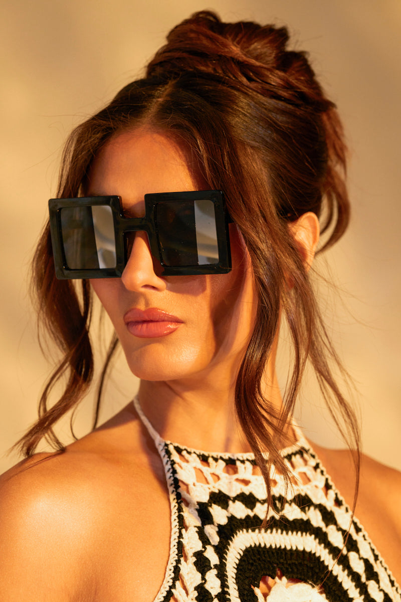 A woman with long hair in a loose bun wears chic square-shaped REALLY LIKE THAT SUNGLASSES, complementing her black and white crocheted top. Warm lighting highlights her face as loose strands softly frame her features.