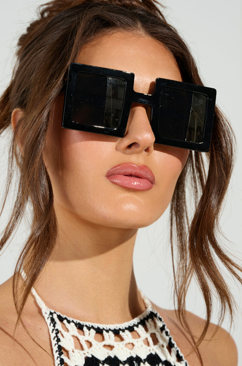 A woman with long brown hair sports oversized square-shaped lenses named REALLY LIKE THAT SUNGLASSES, featuring a half-mirrored design. She pairs these with a crocheted black and white top. Her glossy lips and loose updo stand out against a softly neutral background.