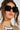 A woman with long brown hair sports oversized square-shaped lenses named REALLY LIKE THAT SUNGLASSES, featuring a half-mirrored design. She pairs these with a crocheted black and white top. Her glossy lips and loose updo stand out against a softly neutral background.