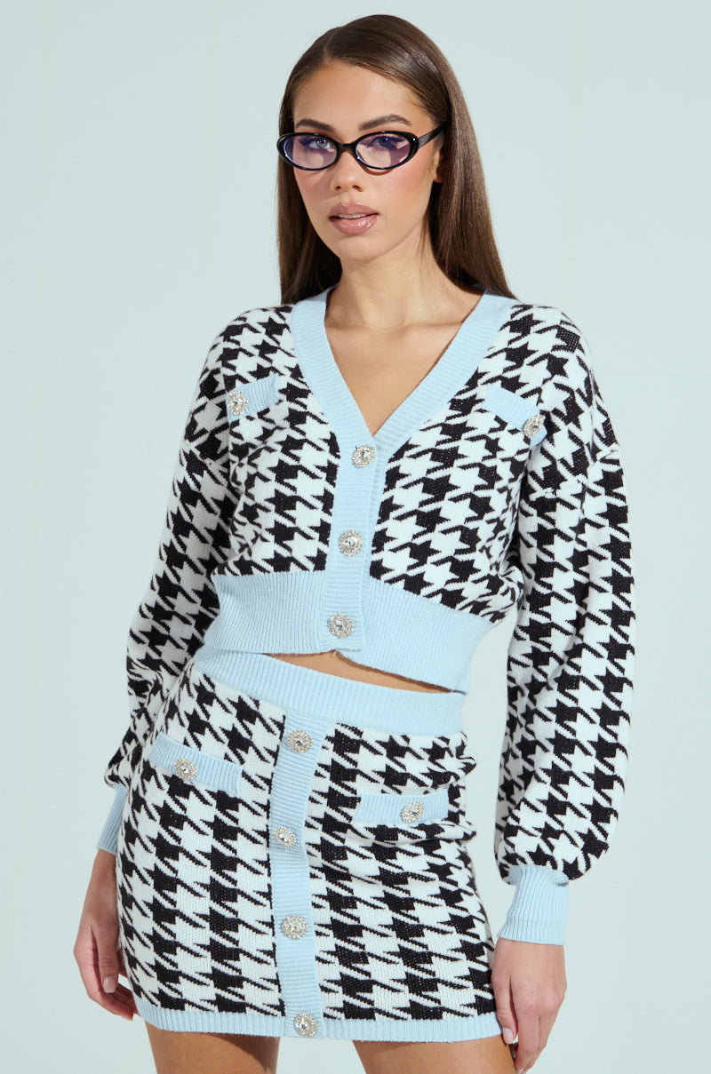 A woman in a preppy outfit wears the black and white PRETTY AND POSH HOUNDSTOOTH CARDIGAN with a matching high-waisted skirt, both featuring light blue trim. Her long brown hair flows under sunglasses, and rhinestone buttons add sparkle to the cardigan.