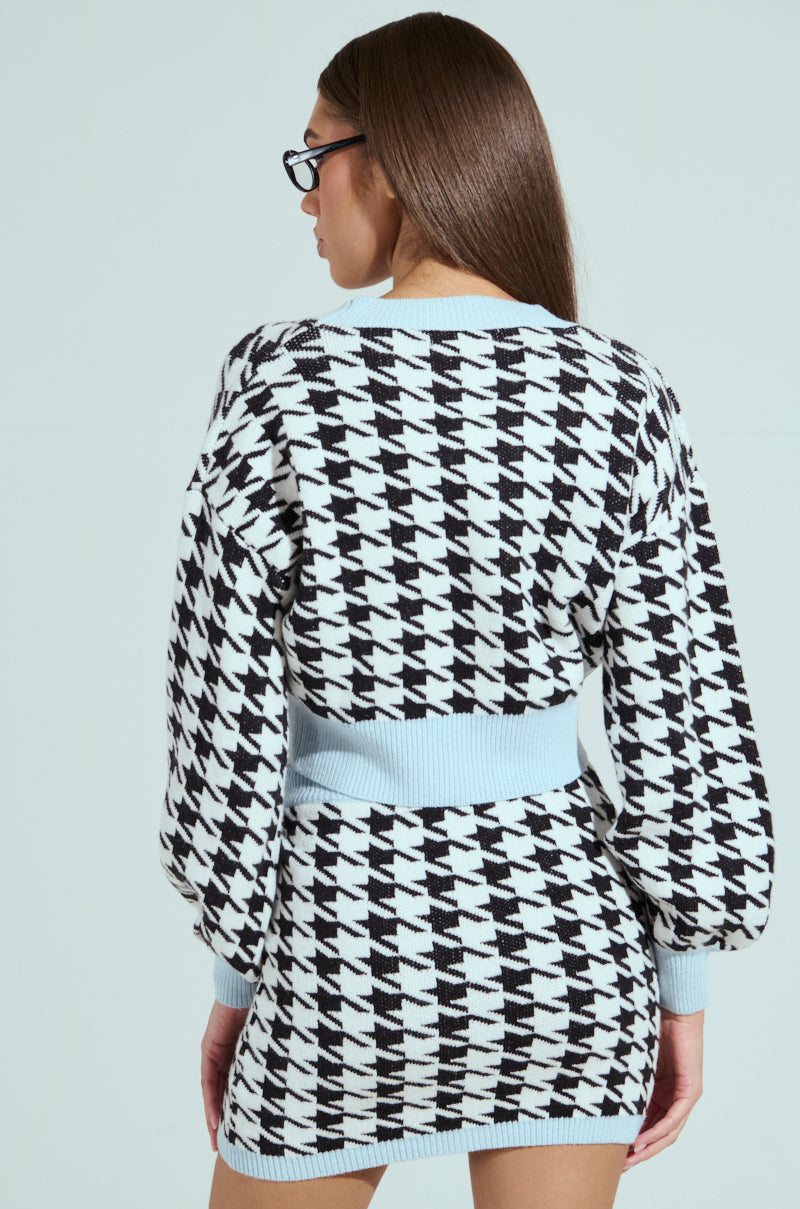 A woman in a black and white PRETTY AND POSH HOUNDSTOOTH CARDIGAN, part of a preppy ensemble, looks away from the camera. She has long brown hair and wears black glasses. The piece, with a fitted waist and balloon sleeves, stands out against the plain light background.