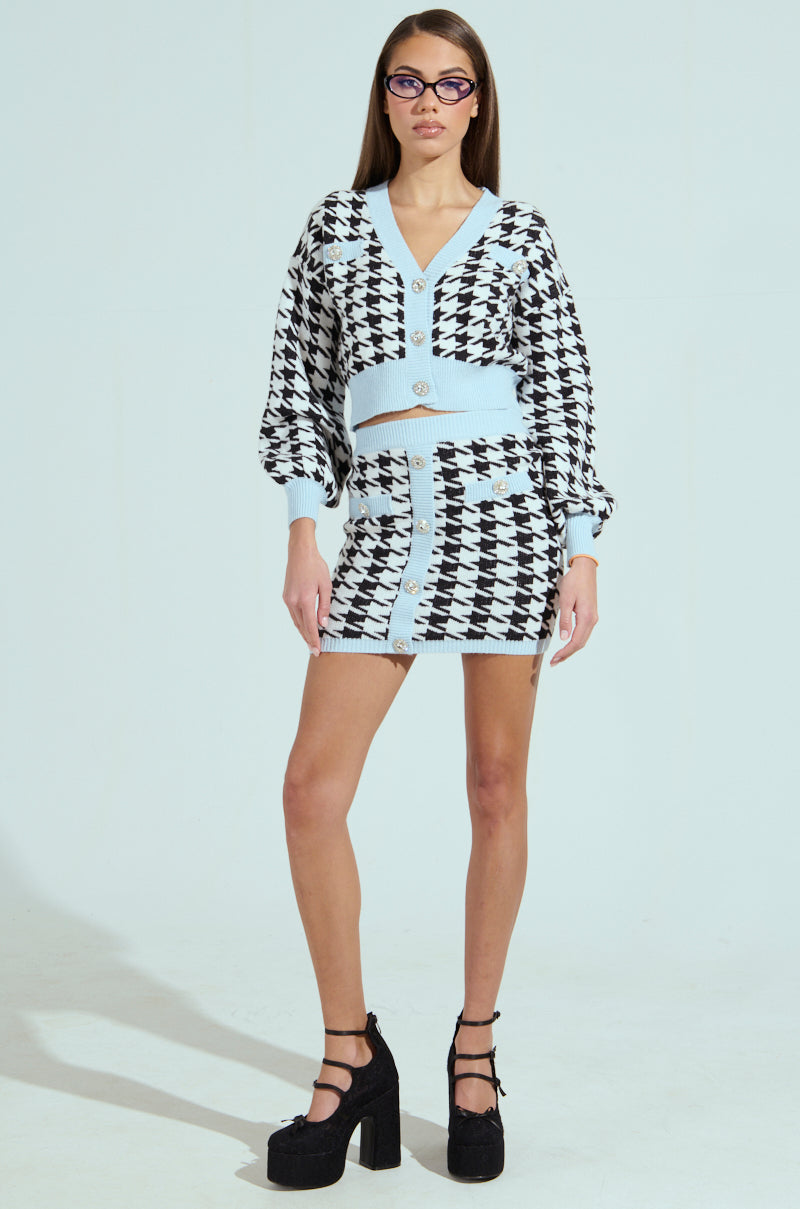 Standing against a plain background, a person wears the PRETTY AND POSH HOUNDSTOOTH CARDIGAN paired with a matching skirt. This ensemble, featuring glimmering rhinestone buttons and light blue accents, is completed with black platform shoes and glasses for a preppy vibe.