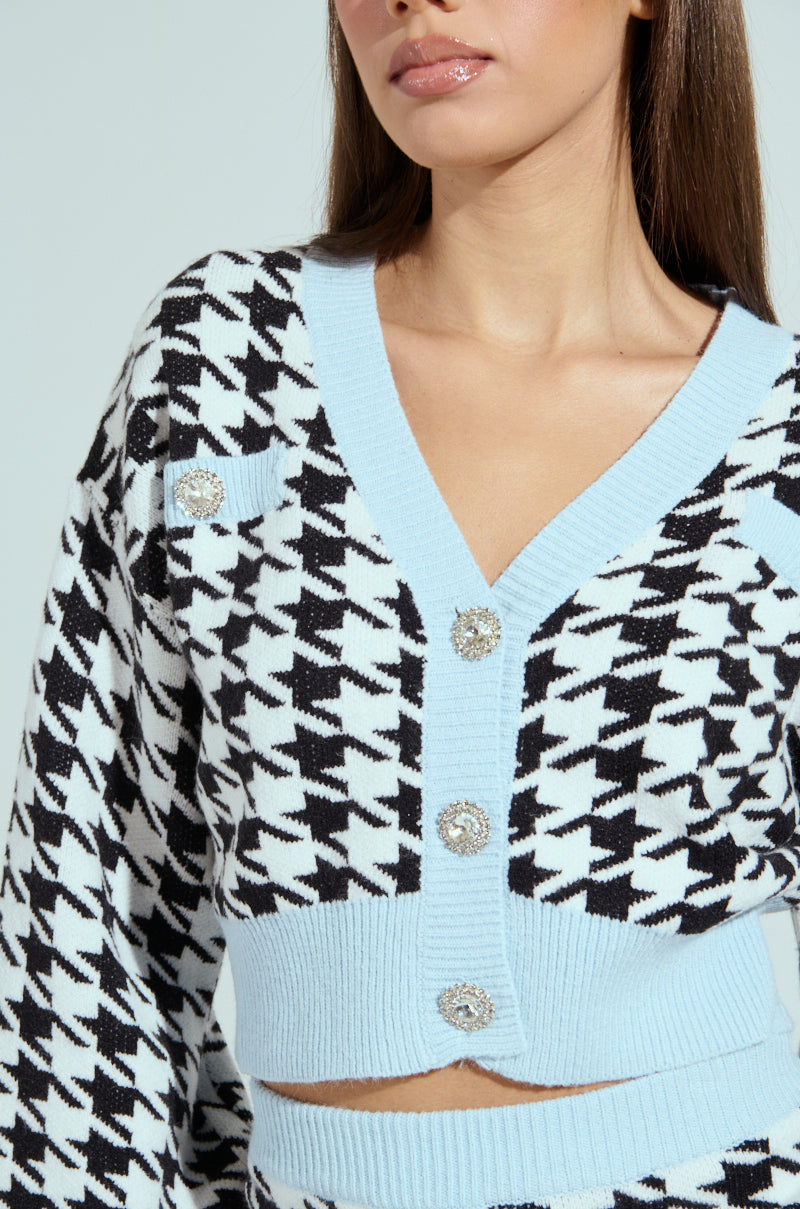 A person in the PRETTY AND POSH HOUNDSTOOTH CARDIGAN, featuring a black and white pattern with rhinestone buttons and light blue trim, pairs it with matching high-waisted bottoms. The chic look enhances their long hair and neutral makeup perfectly.