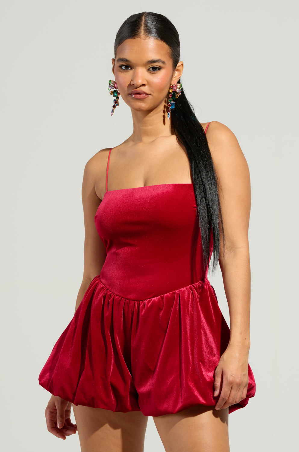 In a stylish holiday outfit, a woman showcases the THIS MOMENT Velvet Romper in red, featuring a sleeveless design with a fitted bodice and flared bubble silhouette. Her long black hair complements her colorful dangling earrings, set against a simple light gray background.