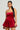 In a stylish holiday outfit, a woman showcases the THIS MOMENT Velvet Romper in red, featuring a sleeveless design with a fitted bodice and flared bubble silhouette. Her long black hair complements her colorful dangling earrings, set against a simple light gray background.