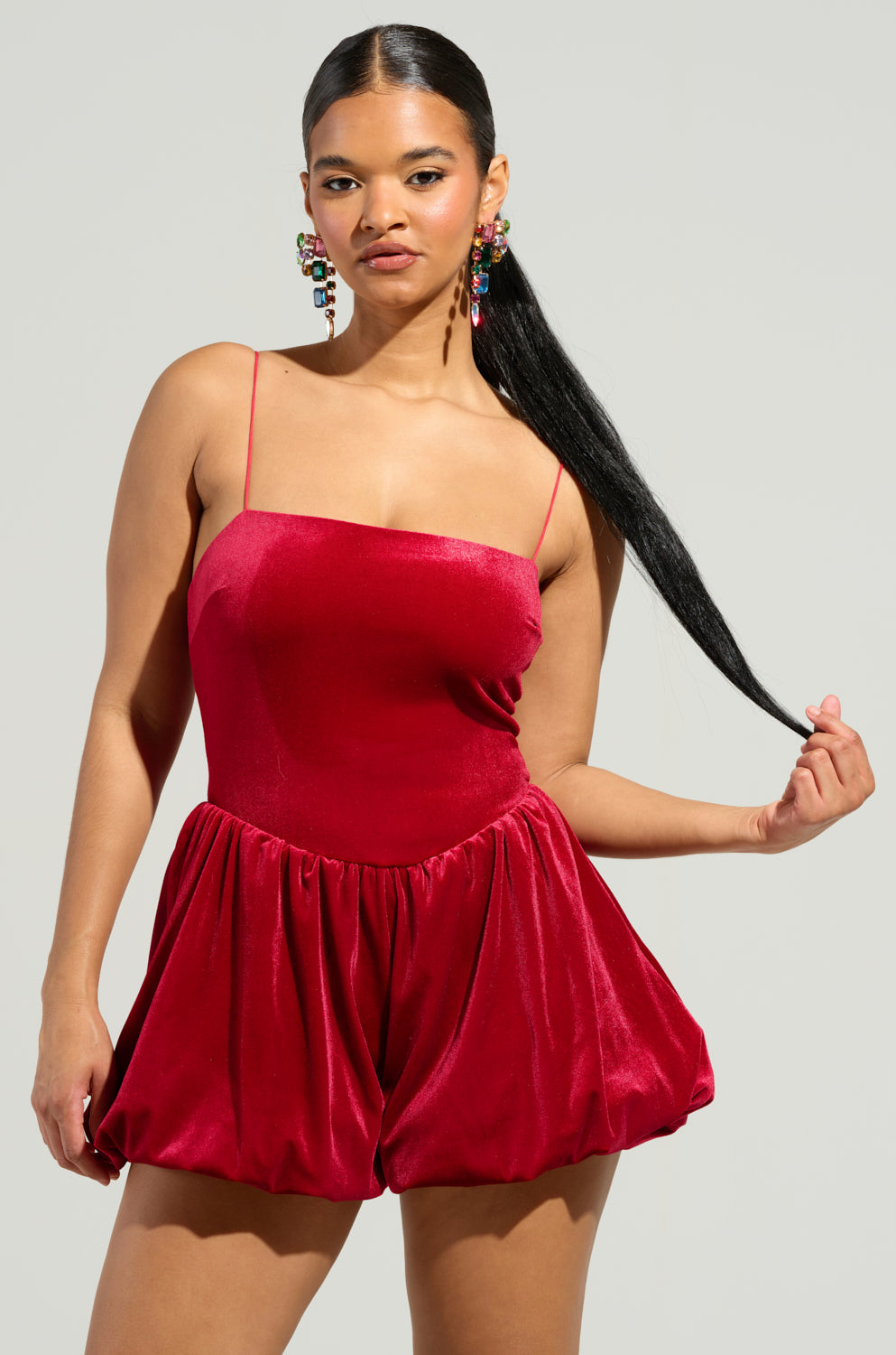 Dressed in the elegant "This Moment Velvet Romper in Red," a woman stands against a simple backdrop, exuding sophistication. The romper's thin straps complement her long black hair that cascades over her shoulder. To enhance her look, she wears vibrant, dangling earrings for added flair. With her left hand gently holding a section of her hair, she gazes confidently at the camera.