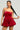 Dressed in the elegant "This Moment Velvet Romper in Red," a woman stands against a simple backdrop, exuding sophistication. The romper's thin straps complement her long black hair that cascades over her shoulder. To enhance her look, she wears vibrant, dangling earrings for added flair. With her left hand gently holding a section of her hair, she gazes confidently at the camera.