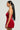 With long black hair styled in a ponytail, they wear a chic holiday ensemble featuring the THIS MOMENT Velvet Romper in Red. Facing to the left, they sport colorful dangling earrings against a plain light gray background.