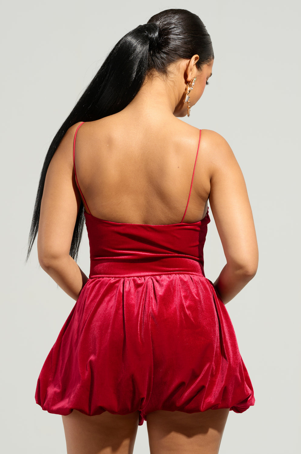 A woman with long black hair styled in a ponytail is wearing the THIS MOMENT VELVET ROMPER IN RED. She is facing away from the camera, accentuating the chic flare of the holiday ensemble's bubble silhouette.