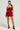 A person stands confidently in the "This Moment Velvet Romper in Red," featuring thin straps and a voluminous flared bubble silhouette. They accessorize with colorful, embellished heels and large decorative earrings against a plain light gray background.