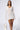 A woman is wearing the CAN'T COVER ME UP CROCHET MINI DRESS, a sheer white mini dress with intricate zigzag patterns, featuring a V-neckline and long sleeves. She is carrying a woven white bag and has long, straight hair. The background is plain and light-colored.