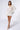 A woman stands against a white background, wearing the CAN'T COVER ME UP CROCHET MINI DRESS over a white swimsuit. She holds a white shoulder bag and wears white high heels. The woman poses with one knee slightly bent and looks straight at the camera.