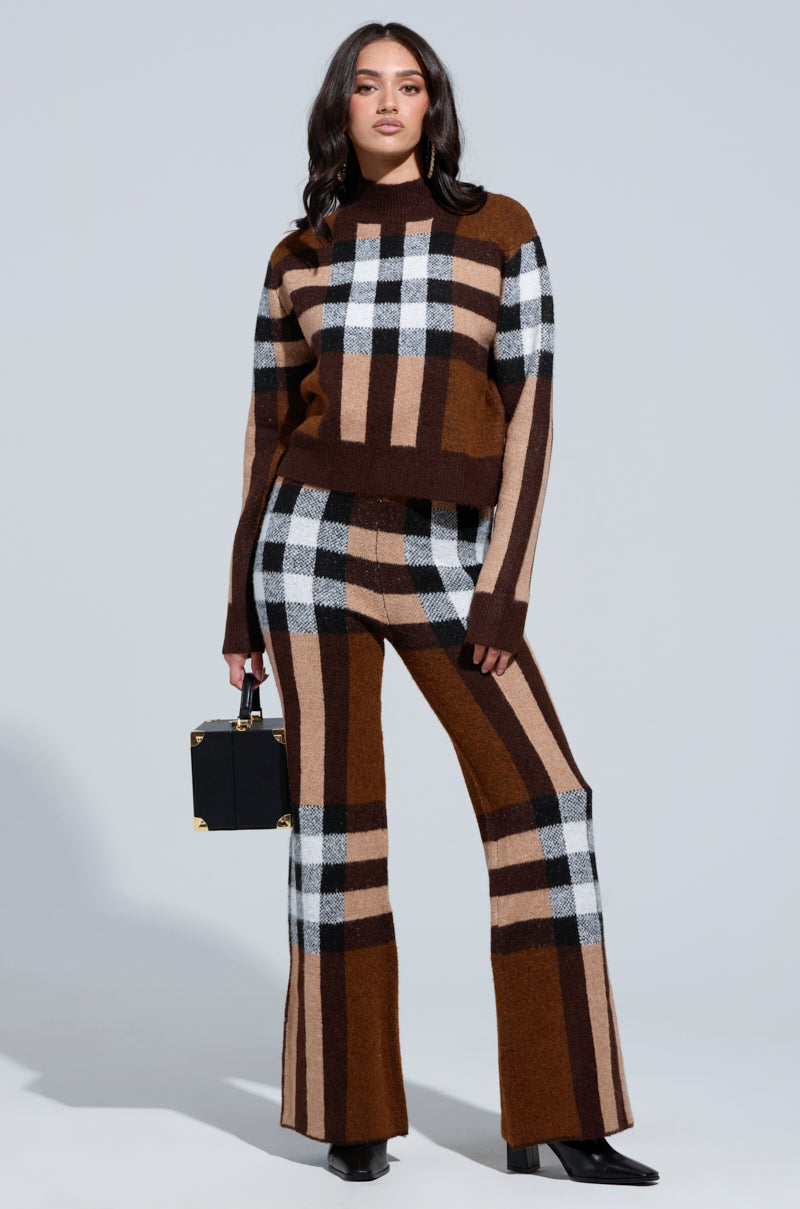 Person wearing the ELISE PLAID KNIT PANT and coordinating sweater, both showcasing bold geometric patterns in brown, black, white, and beige. The outfit features a chic high-rise wide-leg silhouette. They carry a stylish black handbag against a plain, light-colored background. 0
