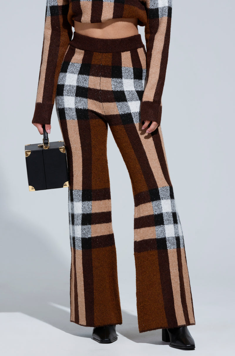 A person in a chic Elise Plaid Knit Pant ensemble stands holding a small black square handbag. This outfit showcases high-rise, wide-leg silhouette pants paired with a matching cropped top with long sleeves. Black ankle boots complete the look. 1