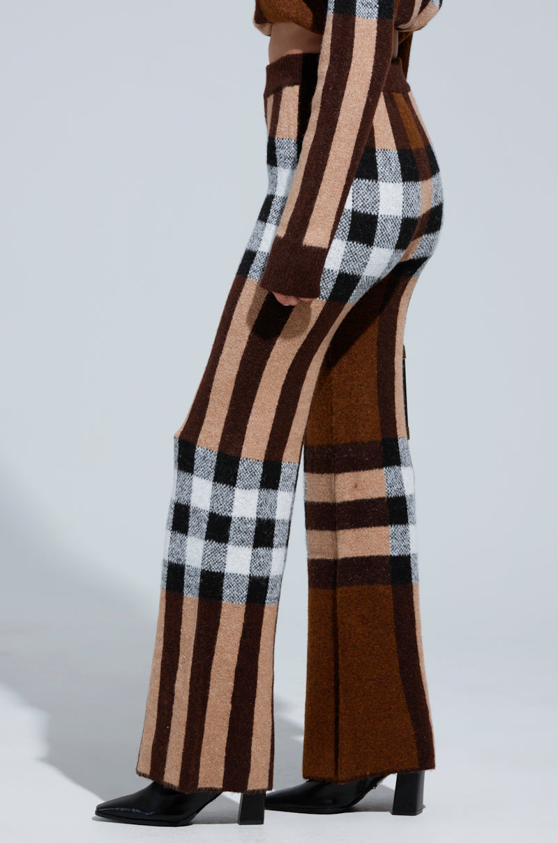 A person showcases a side view of the ELISE PLAID KNIT PANT, stylishly combining shades of brown, black, and beige. The high-rise style boasts a wide-leg silhouette paired seamlessly with the model's black ankle boots. They stand against a plain light gray backdrop. 2
