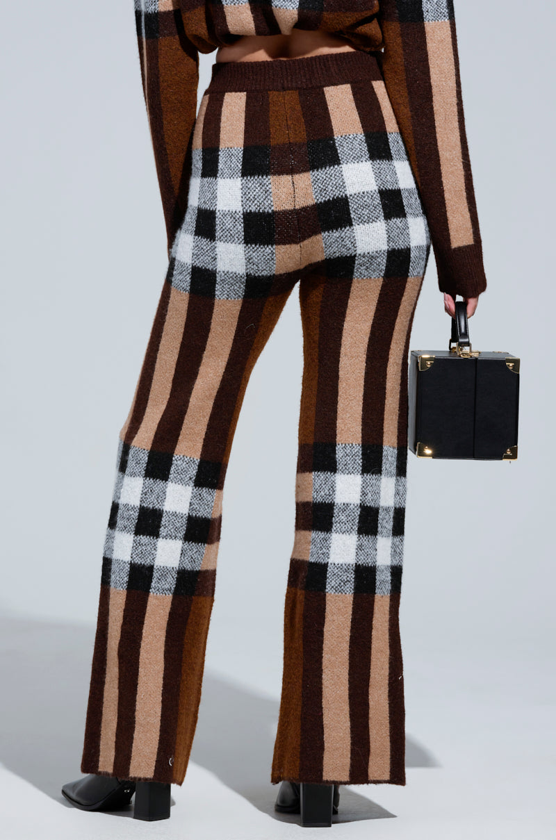 Wearing the trendy ELISE PLAID KNIT PANT, characterized by a high-rise and wide-leg silhouette with brown, black, white, and tan stripes, a person holds a small black square handbag. The simple gray background enhances the stylish look. 3