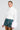A person wearing the Sydney Fur Cuff Sweater in ivory and a green plaid skirt poses against a white background. They have slicked-back hair and are wearing earrings.