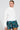A woman is wearing the Sydney Fur Cuff Sweater in ivory paired with a green plaid skirt featuring ruffles. She stands against a plain white background, looking off to the side. Her hair is pulled back, and she has earrings on.