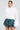 A person poses confidently in the SYDNEY FUR CUFF SWEATER IN IVORY paired with a green plaid ruffled skirt. Bold earrings complete the look, all set against a neutral gray background.