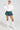 A person exudes confidence in the Sydney Fur Cuff Sweater in Ivory, paired with a green plaid mini skirt and knee-high white boots. They stand against a plain backdrop, highlighting a fashionable and modern ensemble.