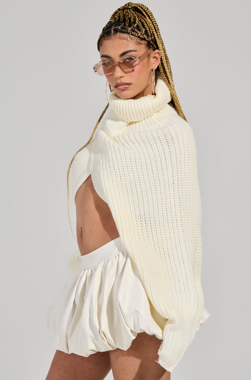 A person wearing sunglasses and large hoop earrings is dressed in the ANNETTA Cropped Long Sleeve Chunky Knit Sweater in cream along with a white, high-waisted skirt. Their hair is styled in long, braided rows and pulled into a high ponytail. This ensemble is perfect for any winter wardrobe against the neutral background.
