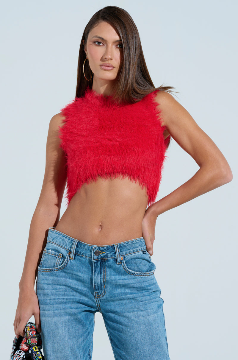 Wearing a SHAGGY RED CROP TOP and blue jeans, the long-haired person confidently stands with one hand on their hip. The faux fur texture pops against the plain light blue backdrop.