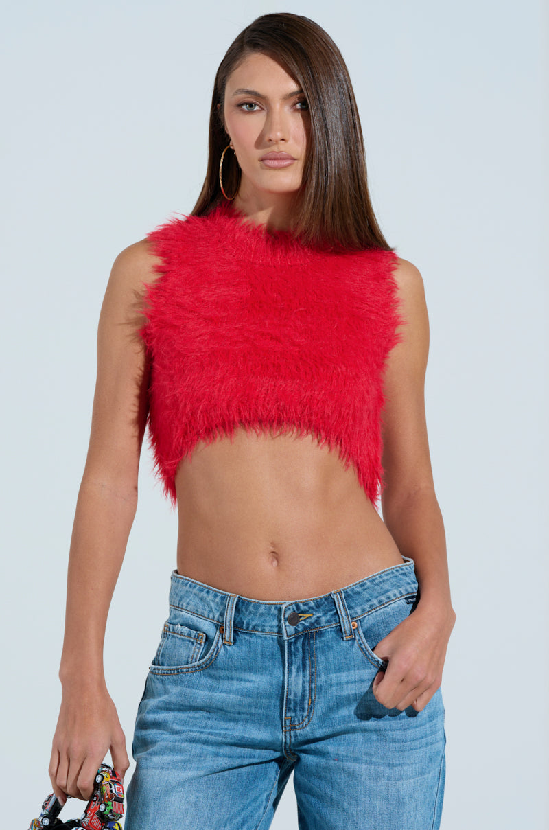 A person with long brown hair poses confidently against a light blue background wearing a SHAGGY RED CROP TOP and blue jeans, holding a colorful patterned bag in one hand to showcase their unique winter fashion sense.