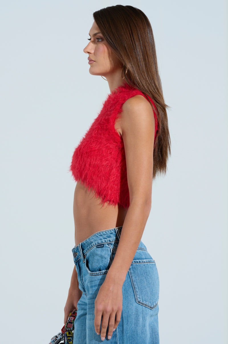 A woman with long brown hair is in profile, displaying winter fashion with a SHAGGY RED CROP TOP and blue jeans against a plain backdrop.