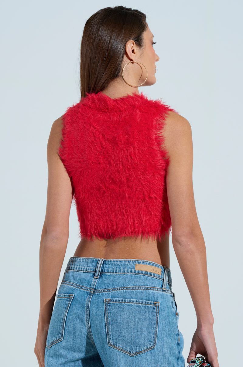 A woman with long brown hair, facing away, flaunts winter chic in a SHAGGY RED CROP TOP and blue jeans. She accessorizes with large hoop earrings and holds a small object against a plain background.