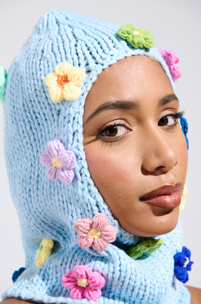 A person wears the BELLA FLOWER BALACLAVA, a cozy knit design with colorful crochet flower appliqués in pink, yellow, and green on a plain backdrop. Their expression is neutral as soft lighting gently illuminates their face. 0