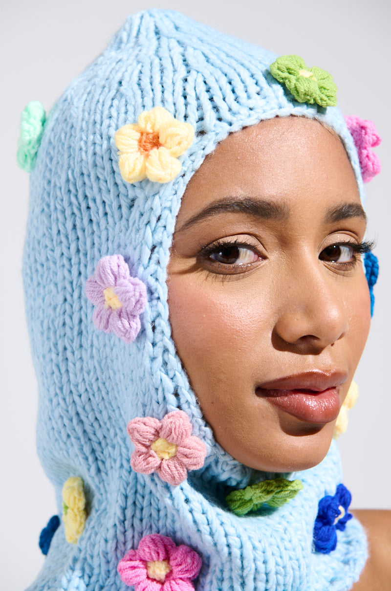 A person dons the BELLA FLOWER BALACLAVA, a light blue knit enhanced with crochet flowers in pink, yellow, purple, and green. The plain backdrop highlights the intricate knit and vibrant hues. 1
