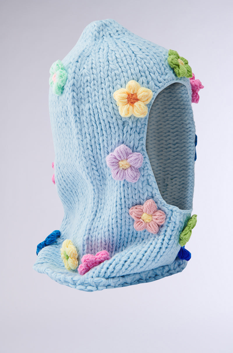 The BELLA FLOWER BALACLAVA is a chunky sky-blue knit featuring vibrant crochet flower appliqués in pink, yellow, green, and purple. It offers a soft embrace with an open face design. 3