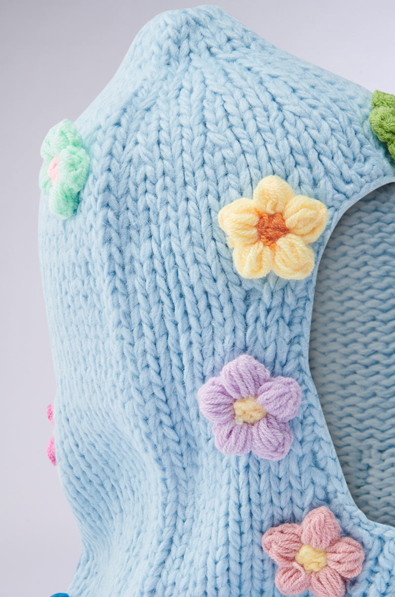 The BELLA FLOWER BALACLAVA features a cozy knit blue fabric with colorful, raised crochet flower appliqués in yellow, pink, and green. Its thick texture suggests warmth for a garment or accessory, showcasing a bright and cheerful aesthetic. 4