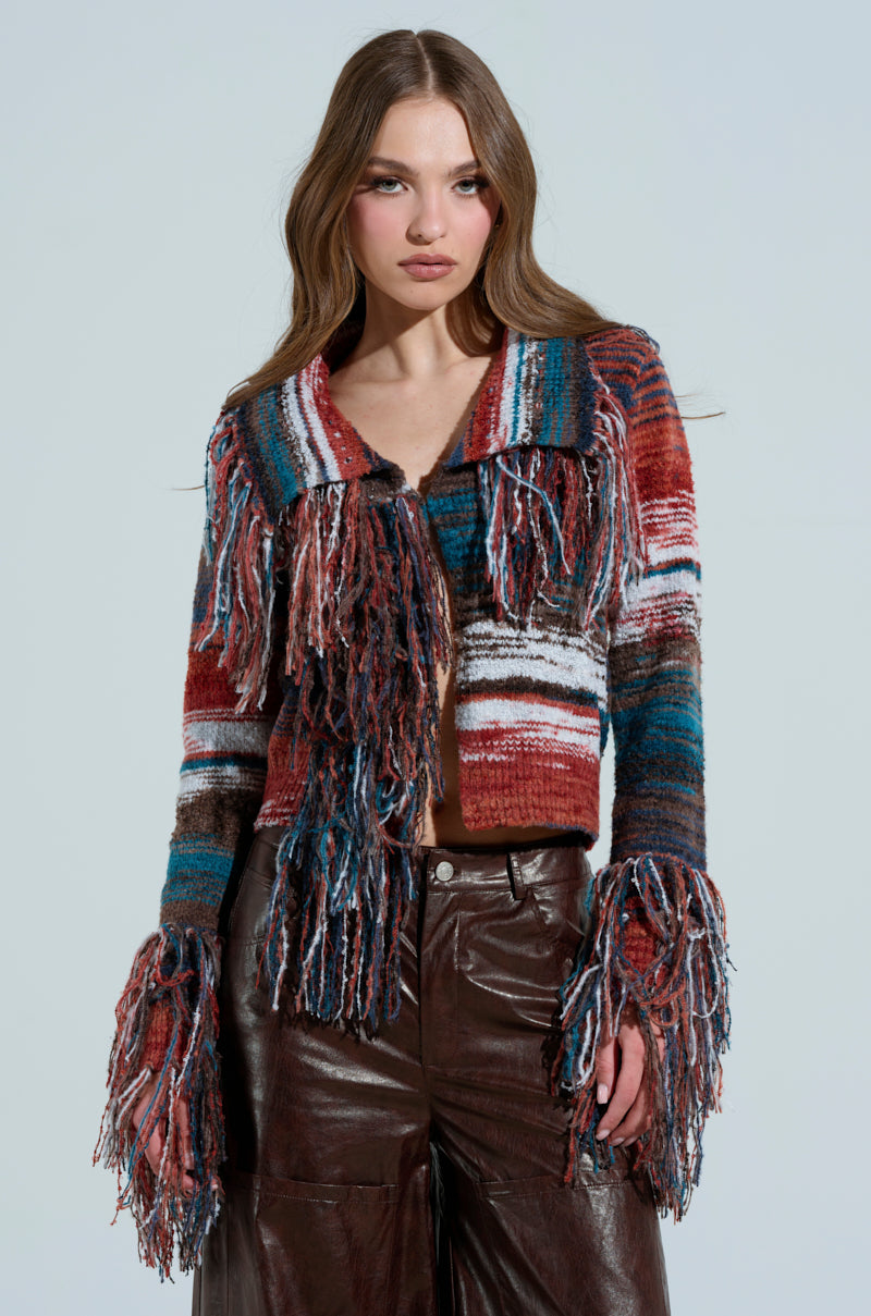 A person wearing a SHAGGY KNIT CARDIGAN with multicolor fringe on the collar and cuffs, paired with brown leather pants against a light gray background.