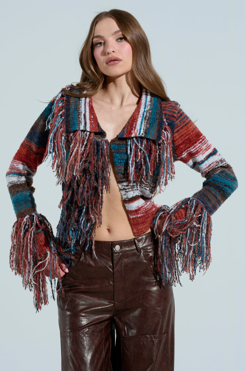 A woman poses in a SHAGGY KNIT CARDIGAN, featuring vibrant red, blue, and white tones. She pairs it with brown leather pants against a light backdrop. Her hands rest on her hips, and her long wavy hair cascades down.