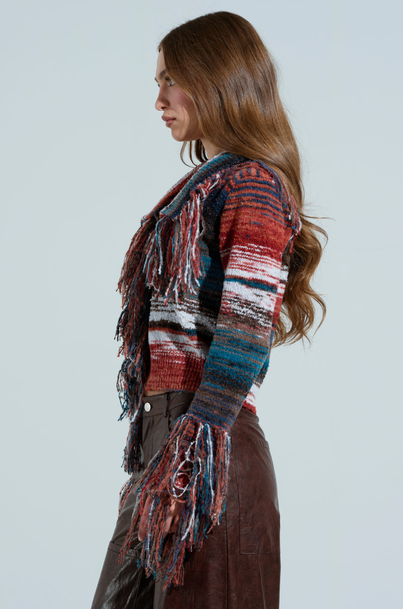 A woman with long brown hair in profile wears a SHAGGY KNIT CARDIGAN featuring fringe trim on the sleeves and front. She pairs it with a brown skirt while standing against a plain gray background.