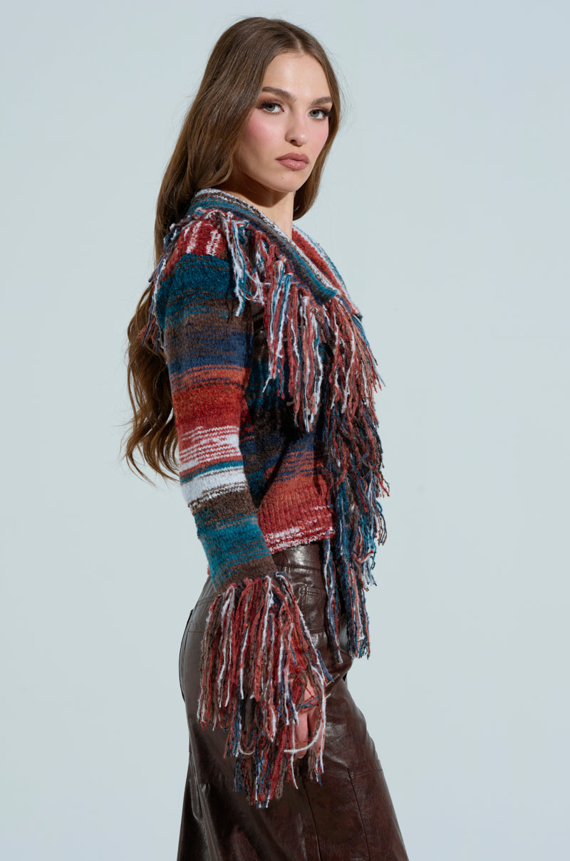 A woman with long brown hair stands in profile against a light background, wearing a colorful SHAGGY KNIT CARDIGAN and brown leather pants. Her confident gaze is directed towards the camera, exuding style.