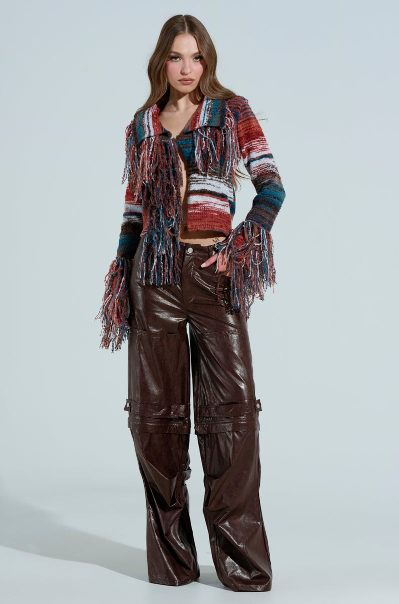A person wearing a SHAGGY KNIT CARDIGAN with fringe trim and dark brown wide-leg leather pants, stands against a light background. They have long, wavy hair and a neutral expression.
