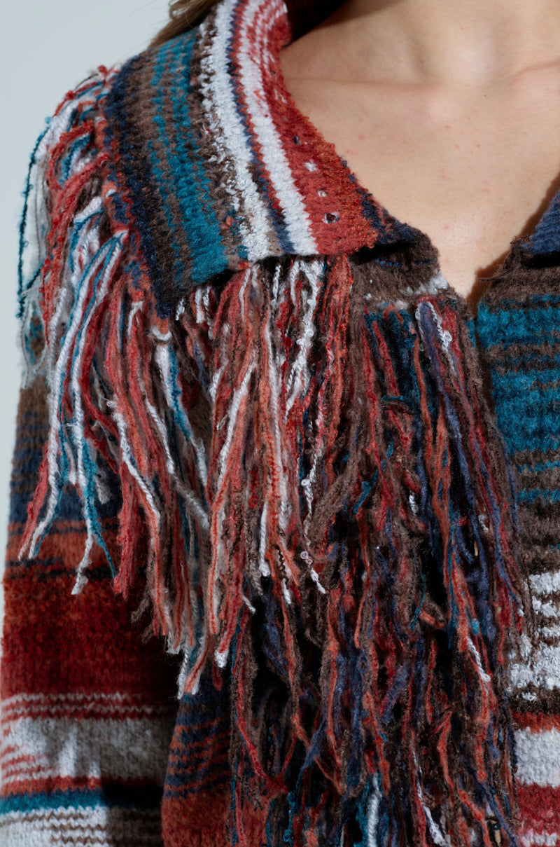 Close-up of someone in a SHAGGY KNIT CARDIGAN with fringe trim, featuring vibrant stripes of red, blue, brown, and white. The textured, fuzzy fabric enhances its shaggy appearance with fringes elegantly draping along the collar and chest.