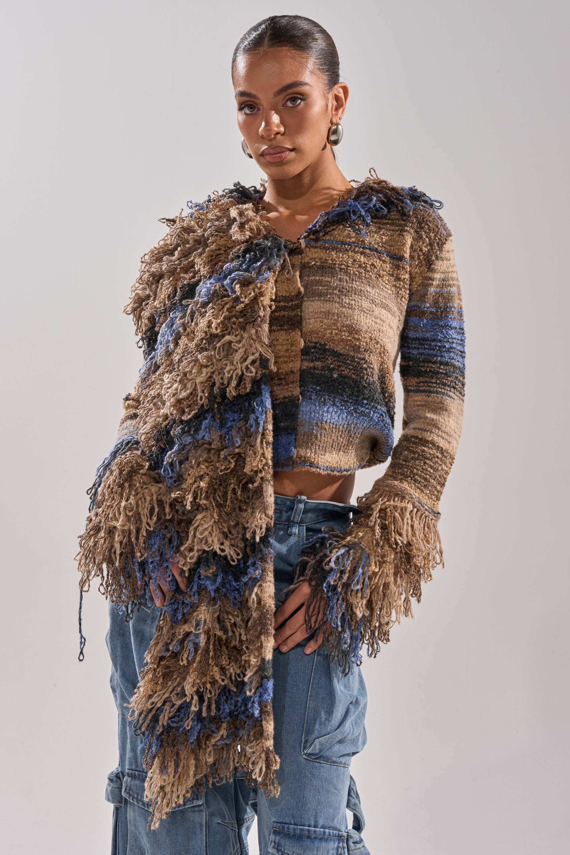 A woman wears the ALWAYS ON MY MIND FRINGE SWEATER—an oversized cropped blue and brown sweater with shaggy fringe on one sleeve and front—paired with loose blue cargo pants, standing against a plain light gray background.