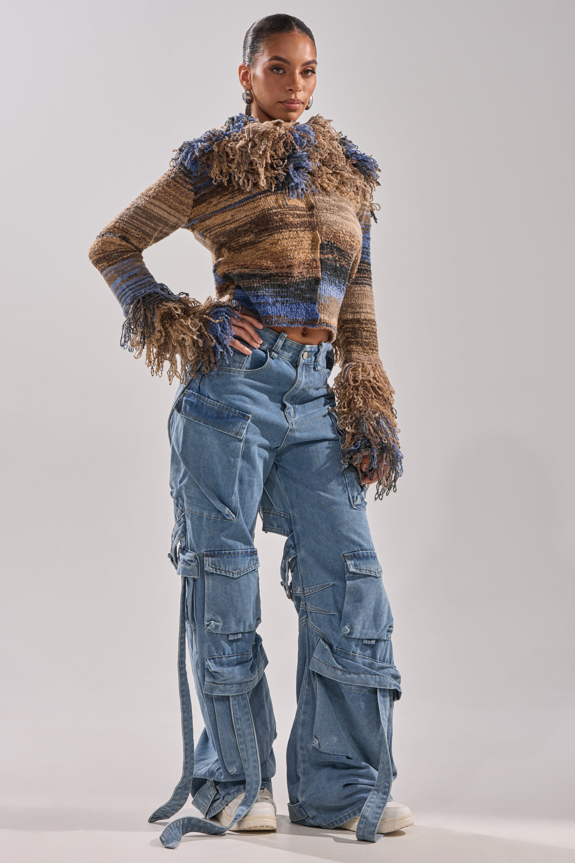 A woman poses in the ALWAYS ON MY MIND FRINGE SWEATER with frayed edges and ombre design, paired with oversized blue cargo jeans featuring multiple pockets and straps, and white sneakers against a plain light background.