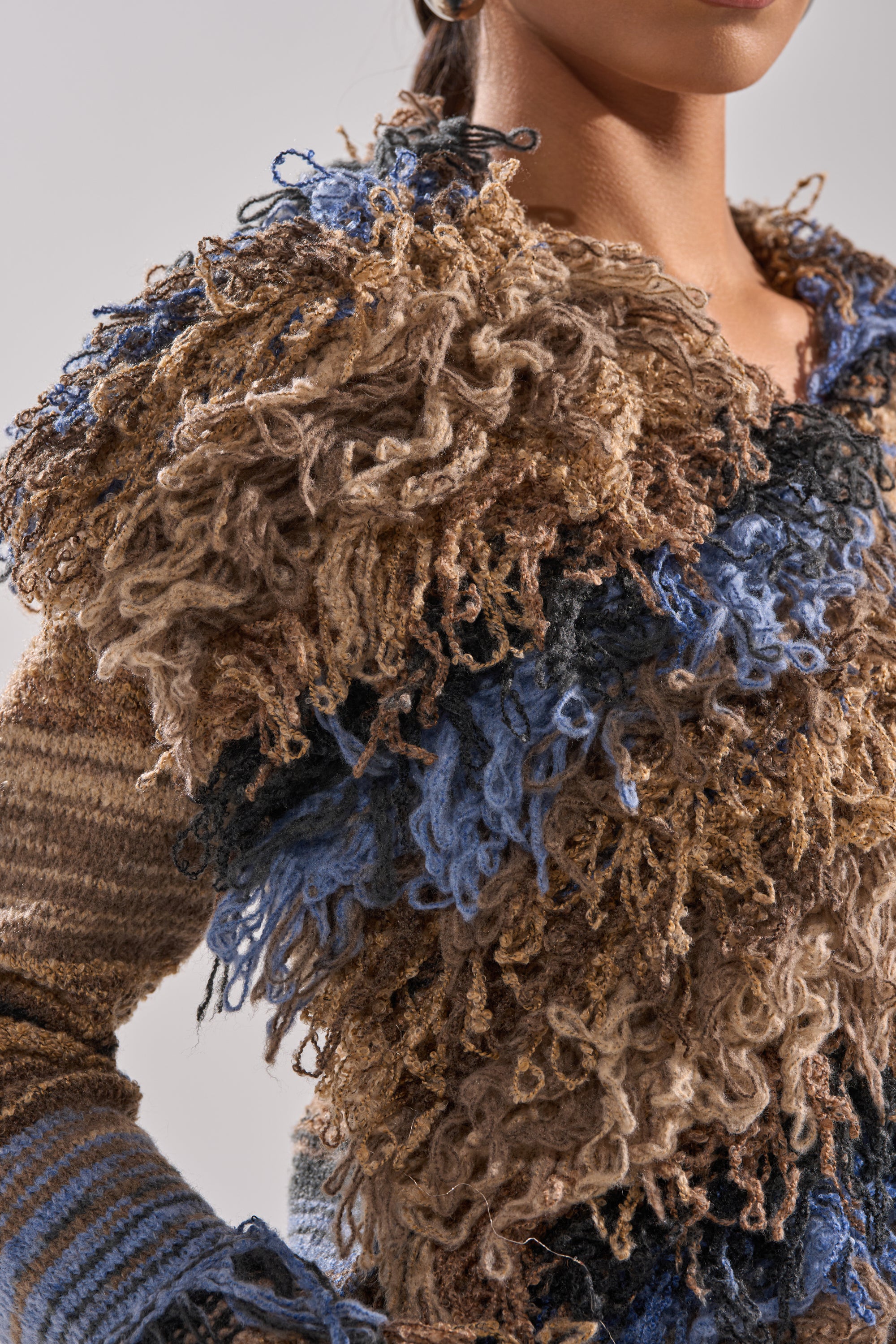 A person wears the ALWAYS ON MY MIND FRINGE SWEATER, an oversized cropped style with thick, curly yarn in brown, beige, blue, and black across the chest and shoulders; their face is partially out of frame.
