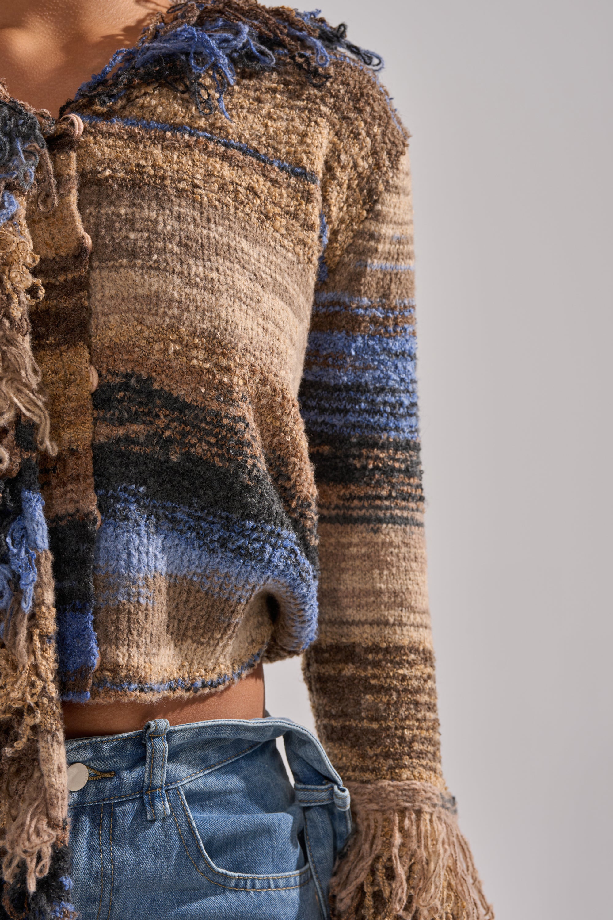 A person wears the ALWAYS ON MY MIND FRINGE SWEATER, a cropped knit with earth-toned and blue ombre stripes, fringe details on the sleeves and hem, styled with high-waisted blue jeans against a plain neutral background.