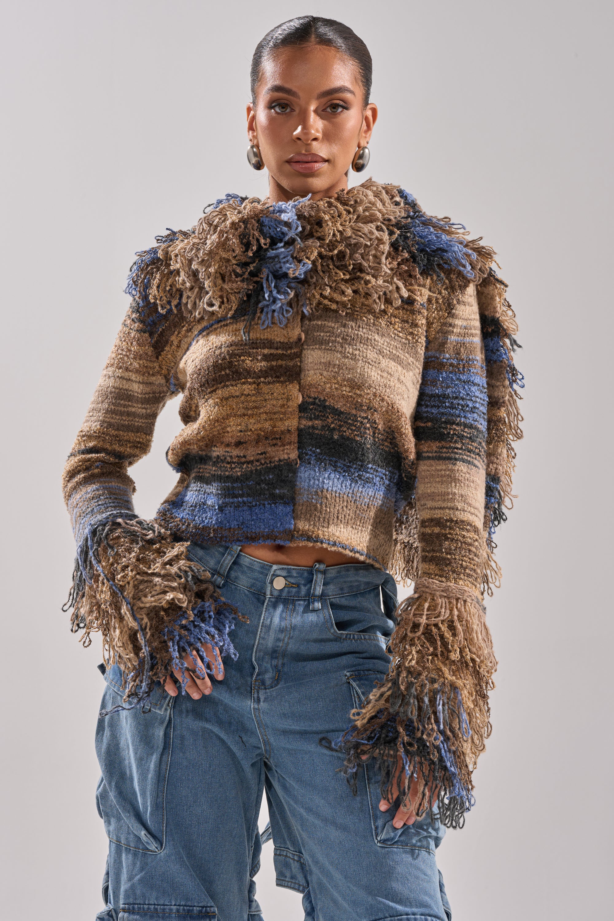 A woman poses confidently in the ALWAYS ON MY MIND FRINGE SWEATER, featuring a brown and blue ombre knit with fringe on the collar and sleeves, paired with high-waisted blue cargo jeans against a plain light background.