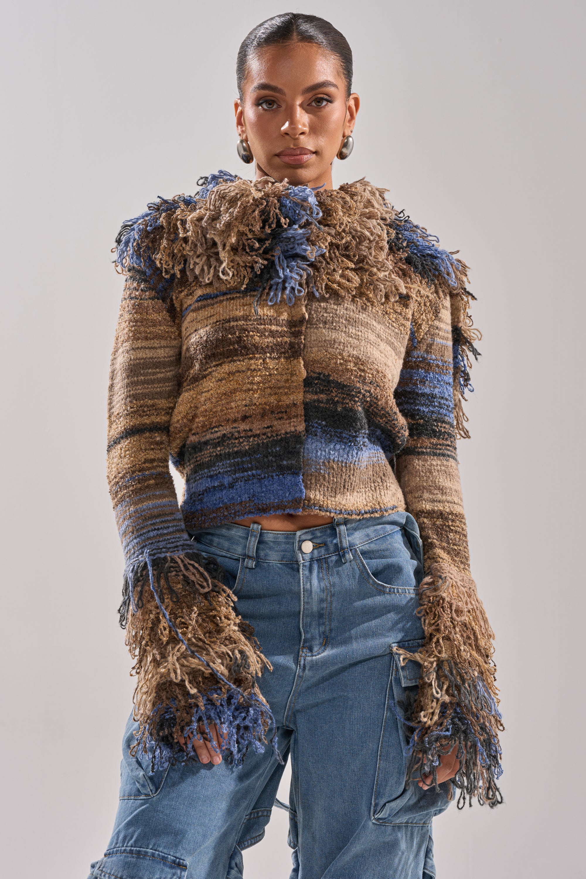 A woman wears the ALWAYS ON MY MIND FRINGE SWEATER, a textured, multicolored brown and blue sweater with shaggy yarn details, paired with high-waisted blue cargo jeans against a plain light background.