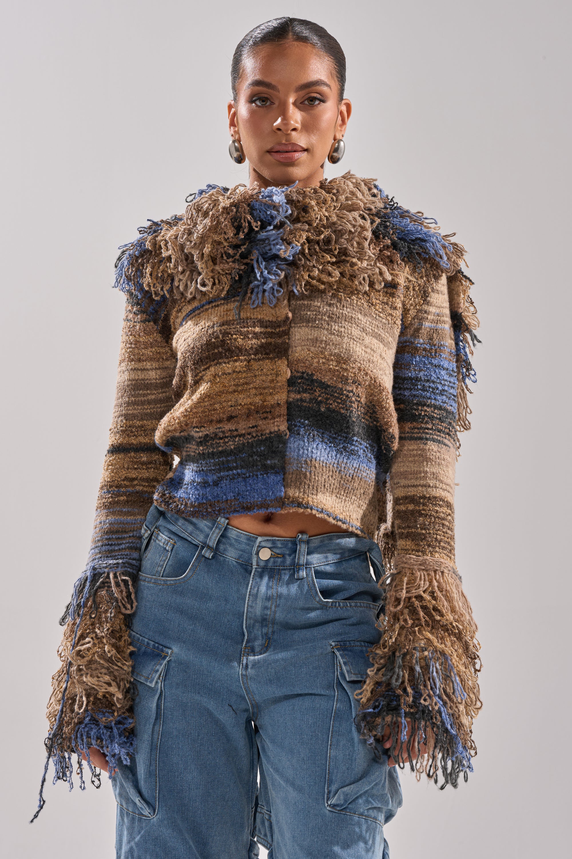 A woman stands against a plain light gray background, wearing blue cargo jeans and the ALWAYS ON MY MIND FRINGE SWEATER—a brown and blue ombre knit with shaggy, fringed details on the collar and sleeves.