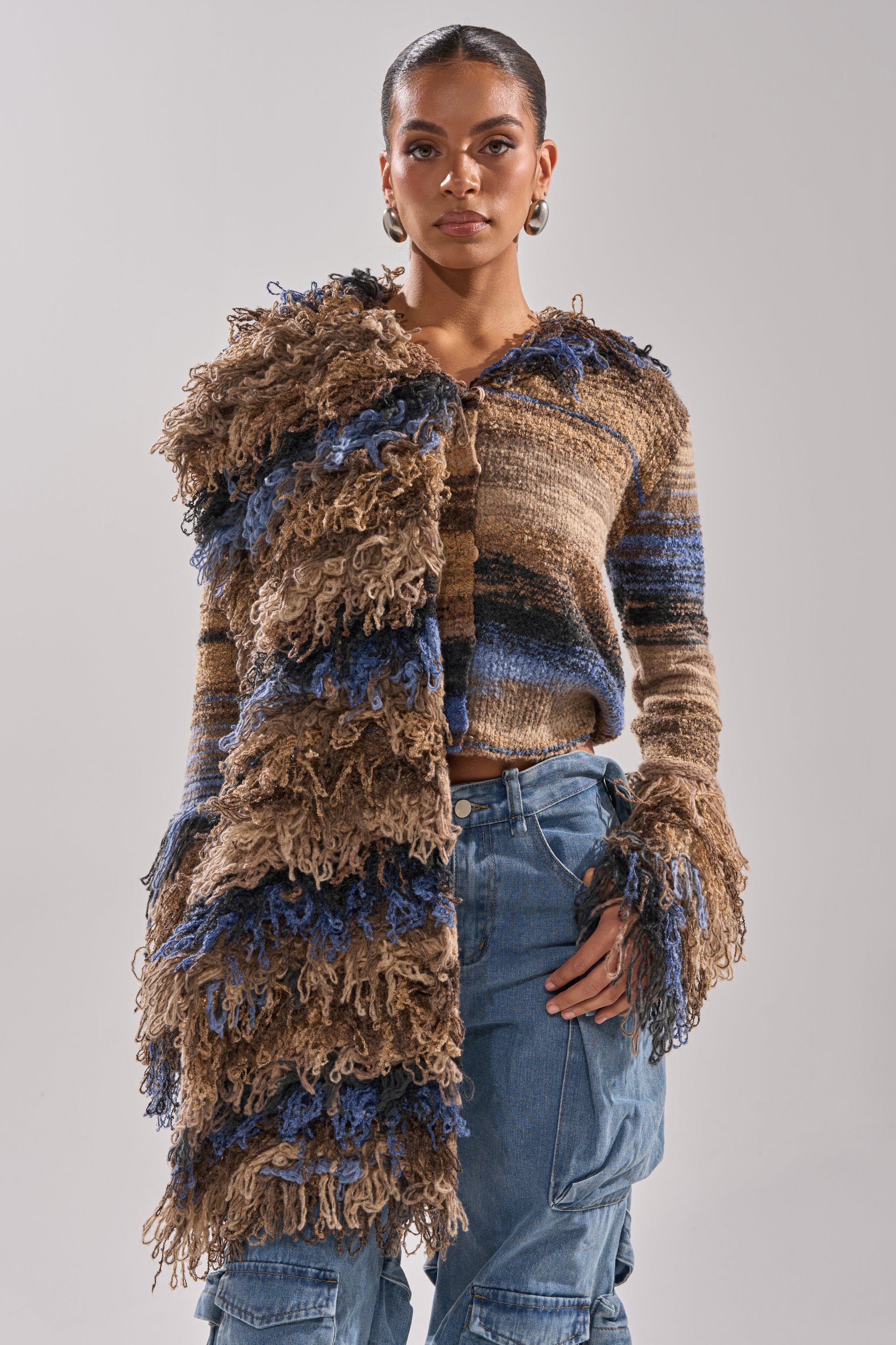 A woman models the ALWAYS ON MY MIND FRINGE SWEATER, a brown and blue ombre striped knit with bold, shaggy fringe on one side and sleeves. She pairs the oversized sweater with blue cargo pants and poses confidently against a plain backdrop.