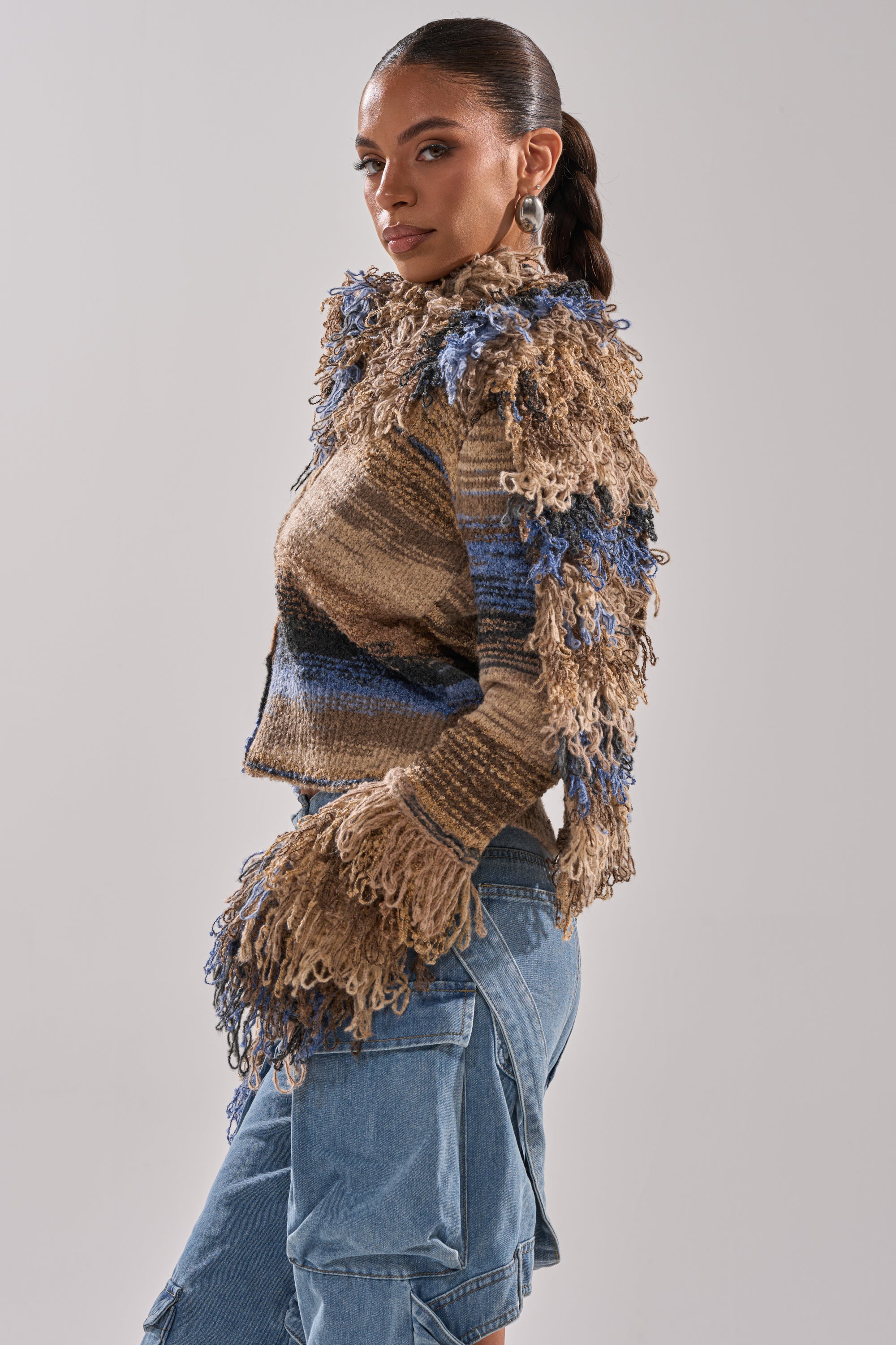 A woman with a sleek ponytail poses sideways in the ALWAYS ON MY MIND FRINGE SWEATER, featuring multi-colored knit textures, paired with light blue cargo jeans against a neutral background.