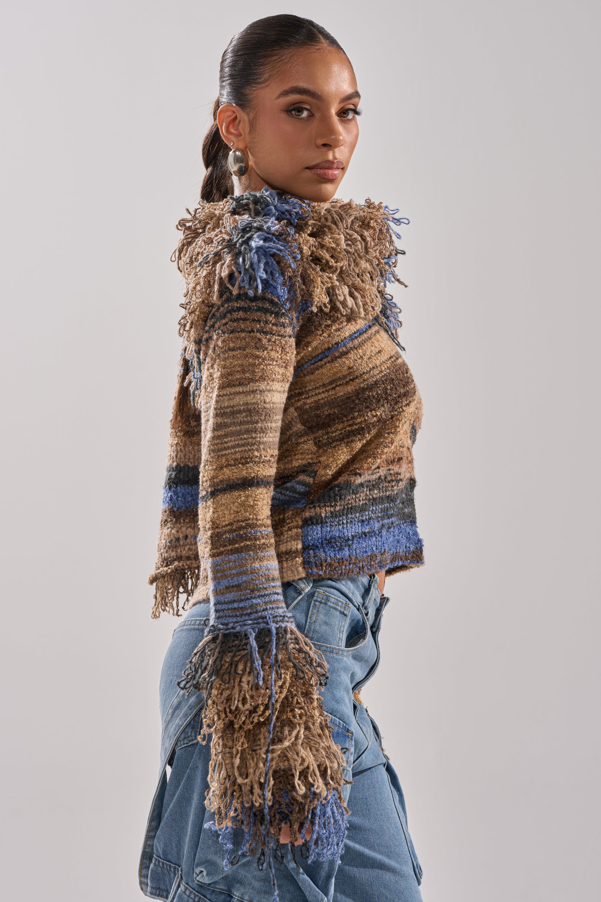 A woman wearing the ALWAYS ON MY MIND FRINGE SWEATER, featuring multicolored brown and blue textures with fringe on the collar and sleeves, pairs it with blue jeans while standing sideways against a light gray background.